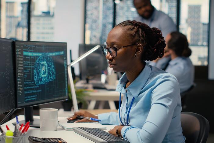 A young woman in a blue shirt works in front of two computer screens.